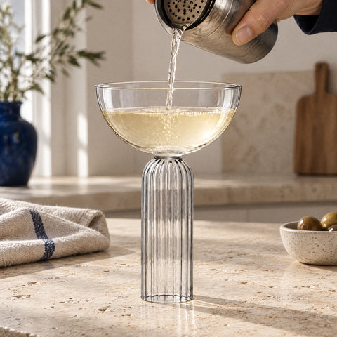 Main versant du shaker un cocktail jaune paille dans la coupe à champagne Torchère, comptoir en travertin et torchon de lin