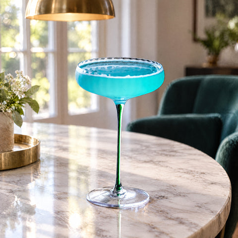 Coupe à champagne Anémone remplie d'un cocktail turquoise, liseré de mousse au buvant, fauteuil en velours flou en fond