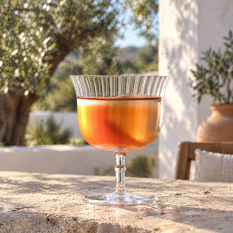Coupe à cocktail Arpeggio remplie d'un cocktail ambré sur une terrasse méditerranéenne, oliviers et murs blancs au loin