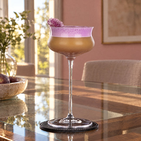 Coupe à cocktail ondulée Volute remplie d'une boisson ambrée à mousse rose, fleur sur le bord, sous-verre en cuir, mur rose