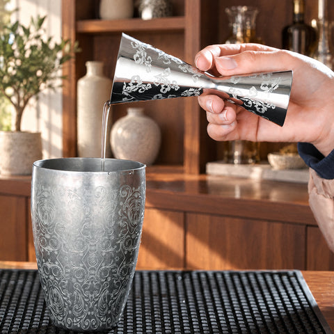 Main versant au doseur Fougère un filet de liquide clair dans une timbale ciselée, meuble de bar en noyer et olivier en pot