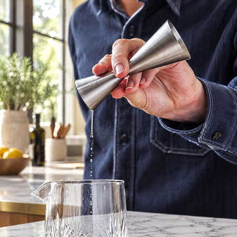 Main de barman versant un filet du doseur Arête dans un verre à mélange en cristal taillé, îlot en marbre blanc