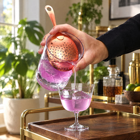 Passoire inox de couleur Palette tenue sur un verre à mélange taillé, cocktail rose versé dans une coupe, chariot de bar