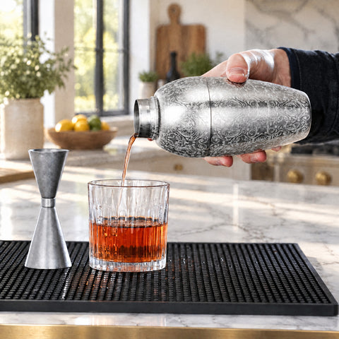 Main versant un filet ambré du shaker à cocktail Rinceau dans un verre bas en cristal, doseur double et îlot de marbre flou