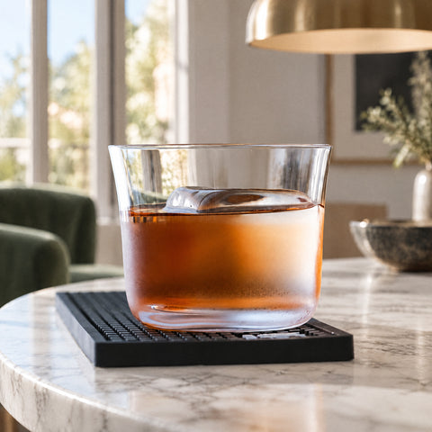 Verre old fashioned Largo embué, cocktail ambré et bloc de glace, sur tapis de bar noir, suspension en laiton floue
