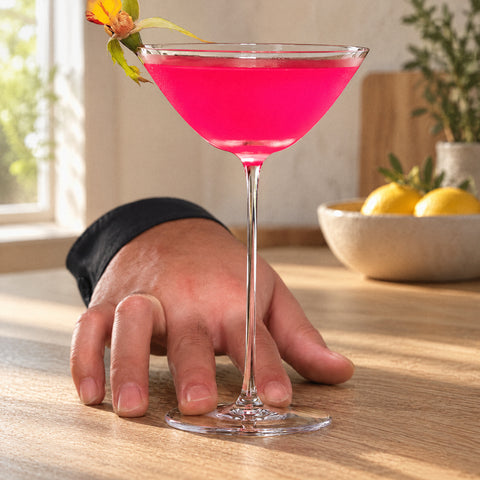 Main posée à plat derrière le verre à martini évasé Crescendo au cocktail rose, fleur orangée au bord, îlot en chêne clair
