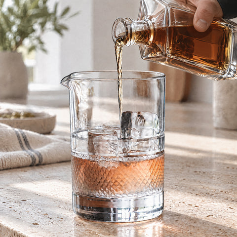 Carafe en cristal versant un spiritueux ambré dans le verre à mélange Jonc garni de deux glaçons, table en bois