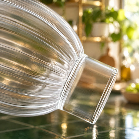 Gros plan sur les côtes verticales du verre à pied bas Dune incliné et sur son socle cylindrique lisse, plan carrelé vert, plantes floues.