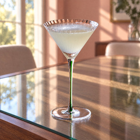 Verre à pied Tourmaline rempli d'un cocktail blanc trouble, mousse en surface, reflet sur le bois, mur rose poudré en fond