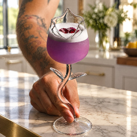 Bras tatoué tenant par la tige le verre à pied en forme de rose Églantine, cocktail violet mousseux, îlot en marbre blanc
