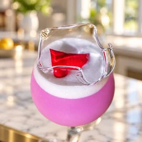 Gros plan sur le bord en pétales du verre en forme de rose, cocktail rose à mousse blanche et pétale rouge, comptoir en marbre.