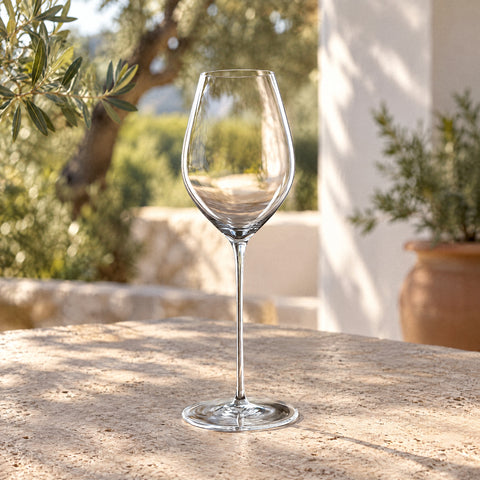 Verre à vin Sépale vide posé sur une table de terrasse méditerranéenne, oliviers et murs blancs flous en arrière-plan
