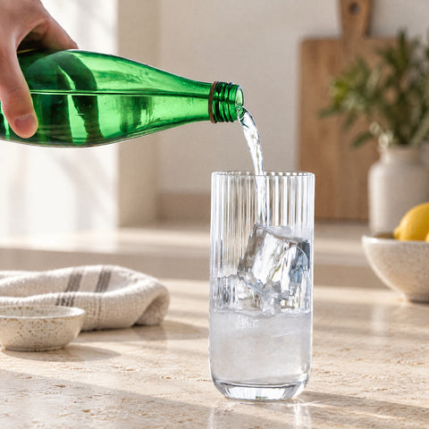 Eau gazeuse versée à la main d'une bouteille verte dans le verre long drink cannelé Arpeggio garni de glaçons, cuisine épurée