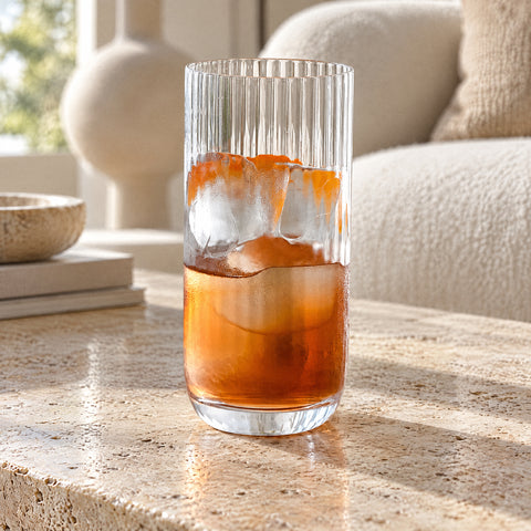 Verre long drink cannelé Arpeggio rempli d'un cocktail ambré avec deux gros glaçons, canapé bouclé crème et vase flous