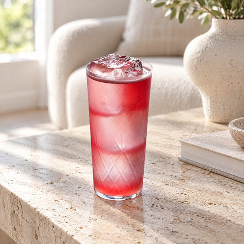Verre long drink Palmette rempli d'une boisson rose sur glaçons, embué, canapé bouclette flou dans un salon lumineux