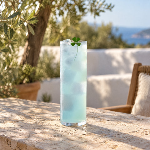 Verre tube Calame rempli d'un long drink bleu pâle glacé, trèfle à quatre feuilles sur le bord, table de terrasse en pierre