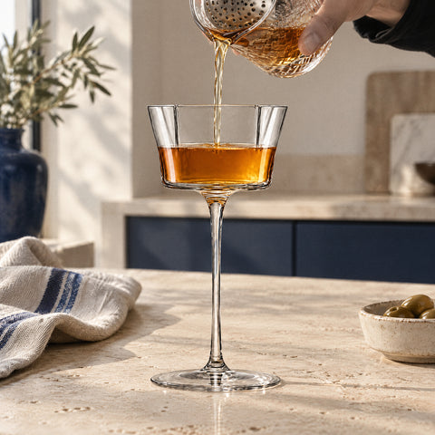 Main versant un mélange ambré orangé à travers une passoire julep dans la coupe à cocktail Quatrefeuille, plan en travertin