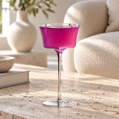 Coupe à cocktail Quatrefeuille remplie d'une boisson magenta, posée sur bois, canapé bouclette et vase en céramique flous