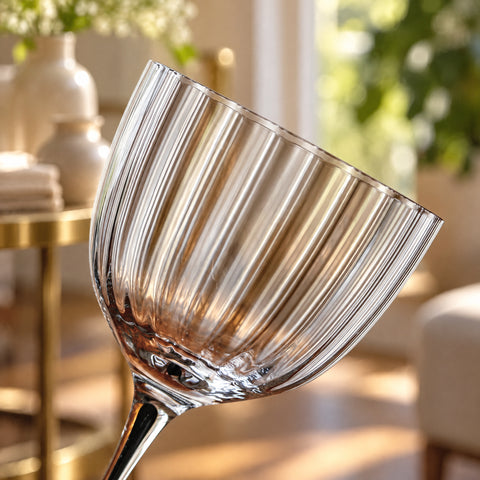 Gros plan sur le buvant côtelé du verre incliné, côtes verticales en relief, tige sombre, vase, plantes et table dorée floues en fond.