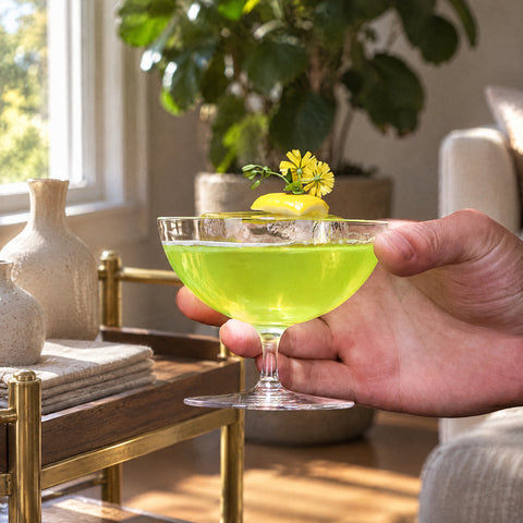 Main soutenant le bol de la coupe à champagne Mailloche, cocktail vert vif, glaçon, citron et fleur jaune, desserte en noyer