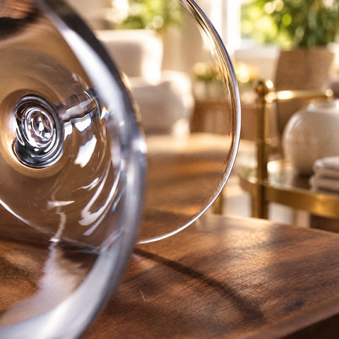 Gros plan sur la coupe à champagne Mailloche couchée sur une table en bois, salon aux fauteuils crème et desserte en laiton flous.