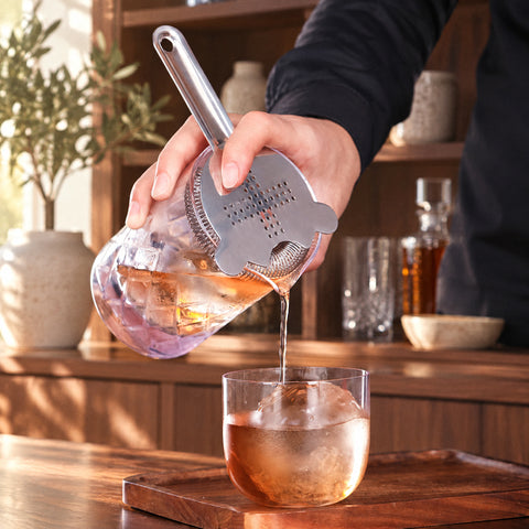 Main filtrant un cocktail ambré au Hawthorne strainer Croisée dans un verre bas à sphère de glace, meuble bar en noyer