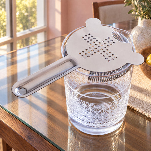 Hawthorne strainer Croisée posé sur un verre à mélange en cristal taillé à motif étoilé, table en bois et mur rose poudré