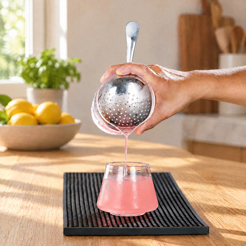 Main pressant le julep strainer Cuilleron sur un verre à mélange, filet de cocktail rose sur glaçons, tapis de bar noir