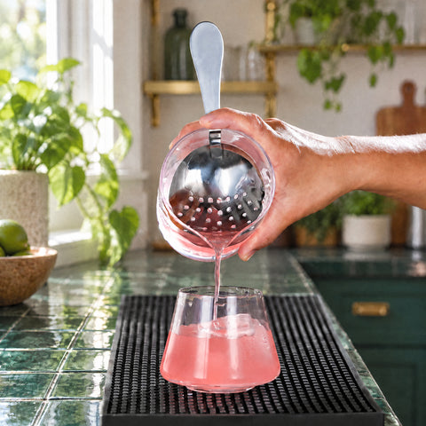 Passoire de barman Peigne dans un verre à mélange tenu en main, cocktail rose versé sur glaçons, tapis de bar noir