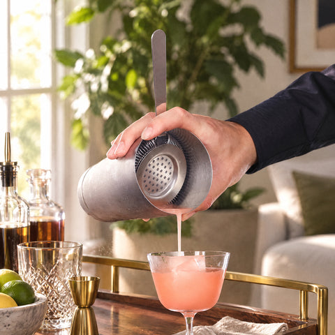 Main filtrant un cocktail rose du shaker par la passoire Hawthorne Vrille dans une coupe, desserte en laiton et noyer