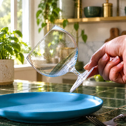 Main tenant par la jambe le petit verre ballon Cabosse incliné, assiette bleue et fourchette, étagères en laiton floues