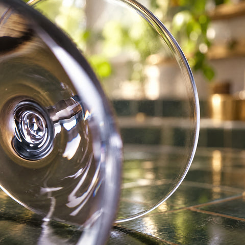 Gros plan sur le petit verre ballon Cabosse couché sur un plan de travail en carreaux verts, plantes et laiton flous en arrière-plan.