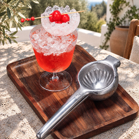 Presse-citron Pressoir ouvert sur une planche en noyer, cocktail rouge à la glace pilée et aux cerises, oliviers flous