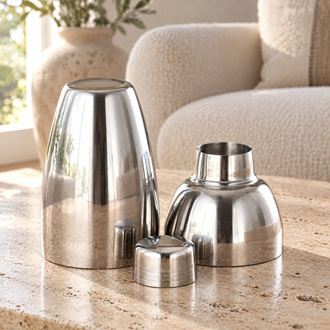 Trois pièces du shaker cobbler Galbe posées sur du bois, corps retourné, dôme filtrant et chapeau, canapé bouclé crème flou