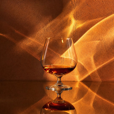 Verre à cognac : Verre à cognac en cristal Verasco rempli de cognac ambré sur une surface miroir, jeu de lumière et caustique dorée, ambiance feutrée