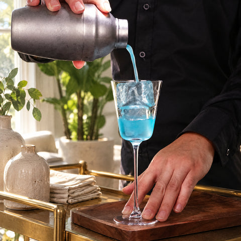 Main versant au shaker un filet bleu dans le verre à cocktail conique à pied Trémie avec deux glaçons, planche en noyer