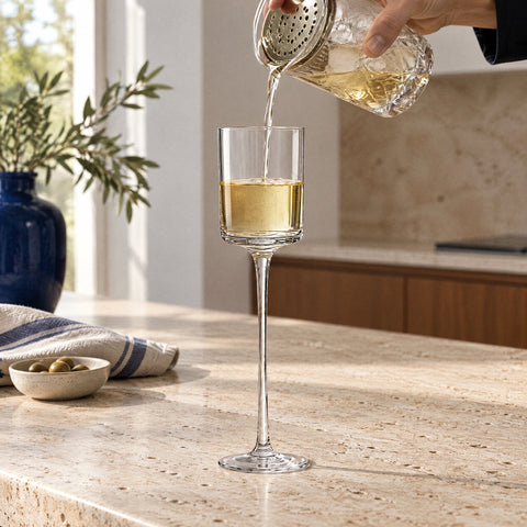 Main versant un mélange doré pâle par une passoire julep dans le verre à cocktail Semplice, comptoir en travertin clair