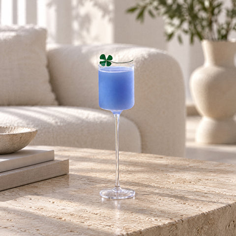 Verre à cocktail Semplice embué, empli d'un mélange bleu pâle, trèfle sur pique en travers du buvant, table en travertin