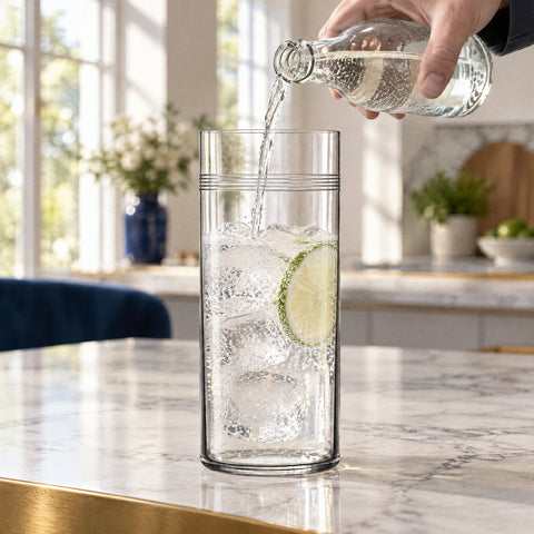 Main versant un gin tonic pétillant dans le verre à cocktail Liseré avec glaçons et citron vert, îlot en marbre blanc