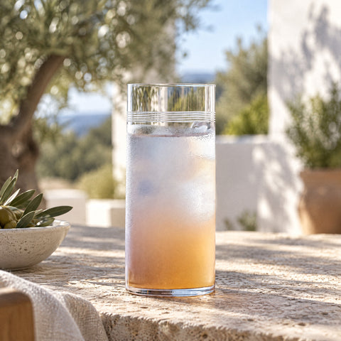 Verre à cocktail Liseré rempli d'une boisson couleur pêche sur glace, tapis de bar noir, oliviers et murs blancs en fond