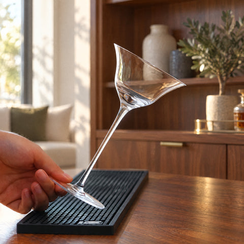 Main tenant par la tige le verre à martini Pavillon incliné sur un tapis de bar, meuble en noyer et olivier en pot au fond