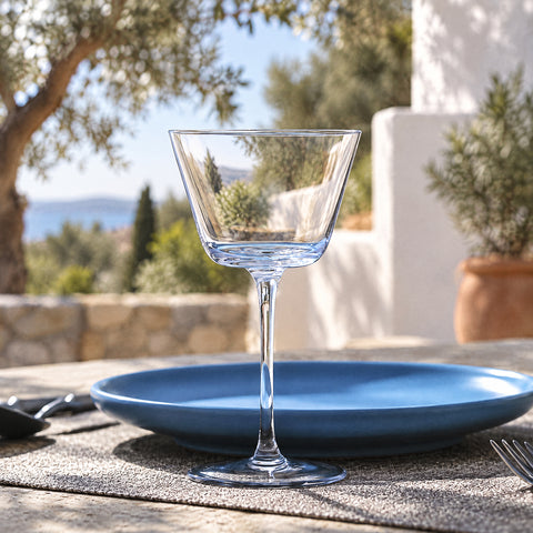 Verre à martini Crescendo vide sur un set tressé, assiette bleu mat et couverts noirs, oliviers et murs blancs en fond