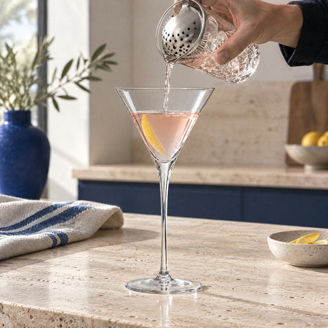Main versant un cocktail rose pâle par une passoire julep dans le verre à martini japonais Épure, comptoir en travertin