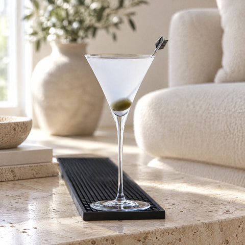 Verre à martini japonais Épure rempli d’un cocktail pâle et trouble, olive verte sur pique en métal, tapis de bar noir