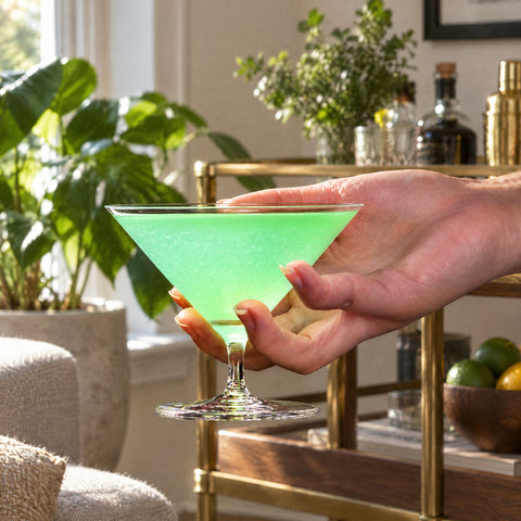 Main soutenant le cône du verre à martini Largo rempli d'un cocktail vert pâle mousseux, desserte en laiton et noyer