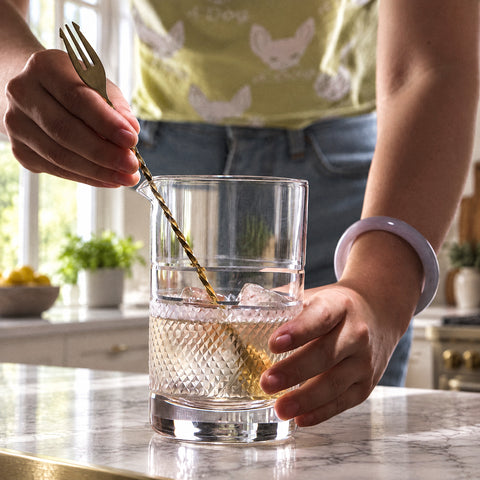 Mains remuant un liquide doré et deux glaçons à la cuillère de bar dans le verre à mélange Jonc, îlot en marbre blanc