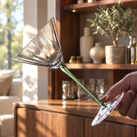 Verre à pied Tourmaline tenu à l'horizontale par une main, meuble de bar en noyer et vases en céramique en fond