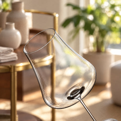 Gros plan sur le calice incliné du verre à vin Nef, panse et départ de tige, desserte dorée, vases et linge plié flous