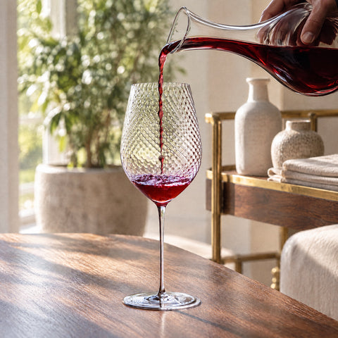Une main verse du vin rouge d'une carafe dans le verre à vin Ténor, au motif optique en relief, sur une table en bois