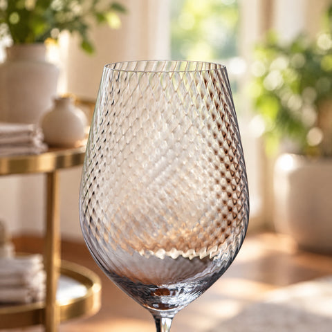 Gros plan sur le calice du verre à vin Ténor, motif optique en relief, desserte en laiton et plantes floues à l'arrière-plan